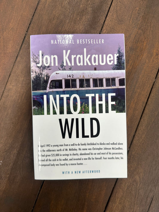 A photograph of the book "Into the Wild" by Jon Krakauer, placed on a wooden surface. The book cover features the title in large white letters, partially overlaying an image of an old, rusted bus with the number "142" visible on its roof. The bus is set against a backdrop of trees and a purple sky. Below the title, a synopsis describes the story of Christopher Johnson McCandless, a young man who hitchhiked to Alaska in 1992, abandoned his possessions, and ventured into the wilderness.